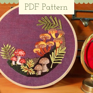 May include: Embroidered mushroom and fern design in a wooden hoop. The design features red and white spotted mushrooms, brown mushrooms, and green ferns on a purple background. The text "PDF Pattern" is at the top.