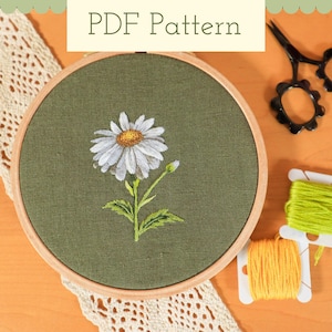 May include: An embroidery PDF pattern showing a white daisy with a yellow centre on green fabric, held in a wooden hoop. Includes black scissors and spools of yellow and green thread. The text "PDF Pattern" is at the top.