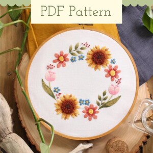 May include: A white embroidery hoop with a floral wreath design. The wreath is made up of pink, yellow, and orange flowers, as well as green leaves. The background is a white fabric.