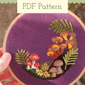 May include: Embroidered mushroom design on a purple fabric, set in a wooden hoop. The artwork features red and white spotted mushrooms, orange and yellow mushrooms, and green foliage. The text "PDF Pattern" is at the top.