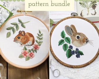 May include: Two circular wooden embroidery hoops containing white fabric with stitched designs. The left hoop features a brown mouse with pink flowers and green leaves. The right hoop shows a brown rabbit with blackberries and green leaves.