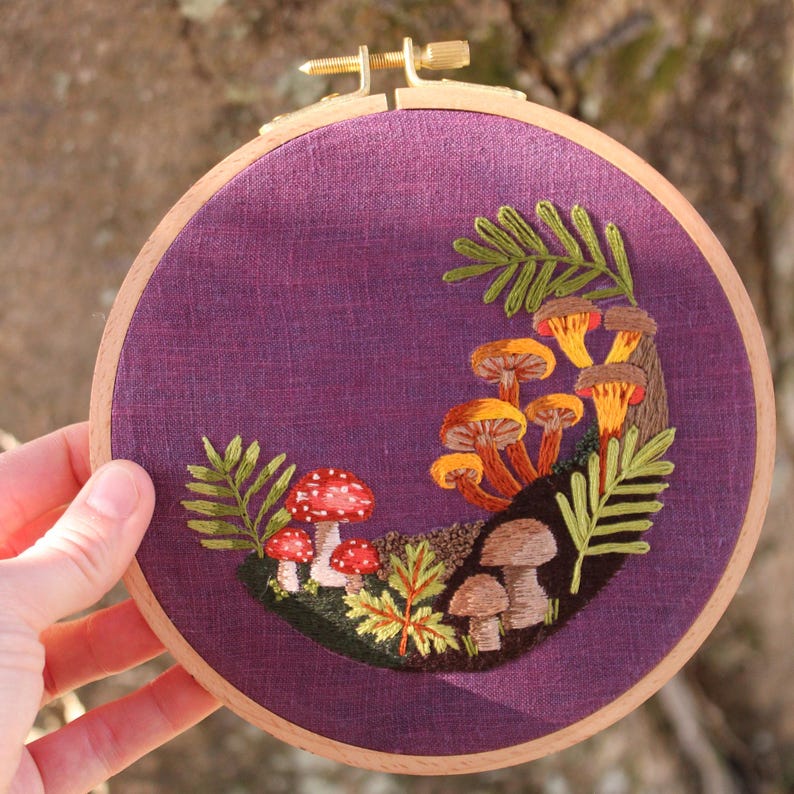 May include: An embroidered artwork featuring a mushroom scene. The design includes various mushrooms in shades of orange, brown, and red, with green foliage. The embroidery is set against a purple fabric background, framed in a wooden hoop.
