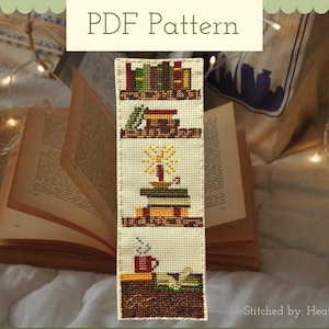 May include: A cross-stitch bookmark depicting bookshelves, a candle, and a coffee cup. The bookmark reads "PDF Pattern" at the top and "Stitched by: Heather" at the bottom. An open book and fairy lights are in the background.