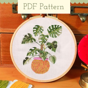 May include: An embroidered plant design in a round hoop. The plant has large green leaves and a yellow and pink pot. The text "PDF Pattern" is at the top. The hoop is set against a wooden background.