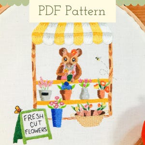 May include: Embroidered hoop art featuring a mouse selling flowers at a market stall. The stall has a yellow and white striped awning and a sign that reads "FRESH CUT FLOWERS". A bee and butterfly are also embroidered.