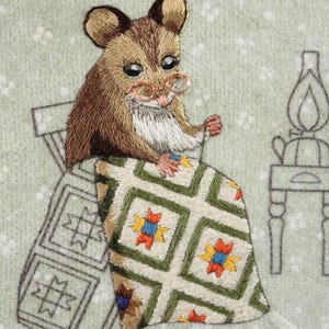 May include: Embroidered image of a mouse wearing spectacles, seated on a chair, and sewing a quilt. The quilt has a green, white, yellow, red, and blue geometric pattern. The background is light green with white dots.