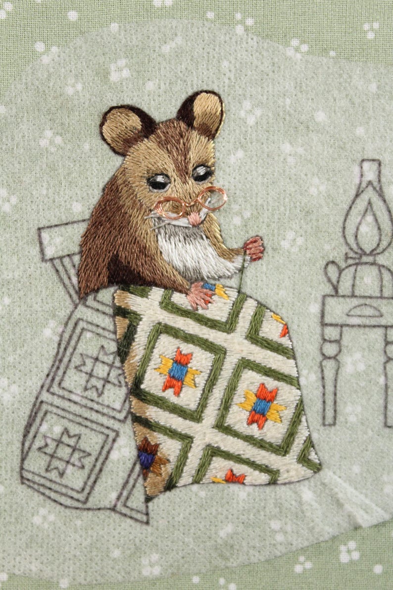 May include: Embroidered image of a mouse wearing spectacles, seated on a chair, and sewing a quilt. The quilt has a green, white, yellow, red, and blue geometric pattern. The background is light green with white dots.
