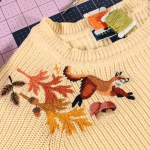 a yellow knit sweater with embroidery along the neckline. The embroidery features a fox leaping over mushrooms, with colorful leaves and acorns