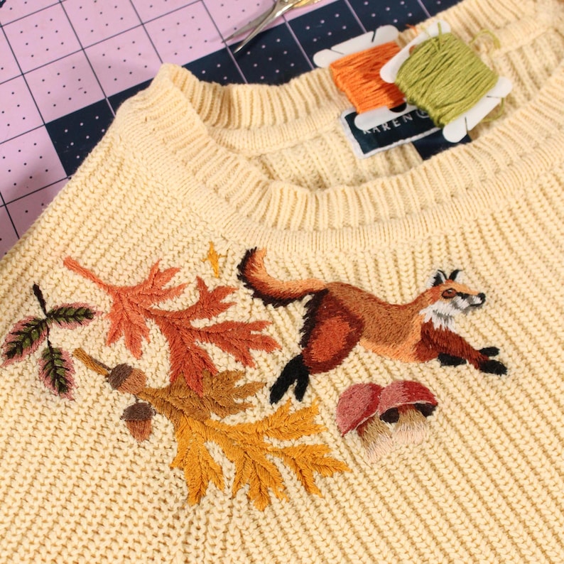 a yellow knit sweater with embroidery along the neckline. The embroidery features a fox leaping over mushrooms, with colorful leaves and acorns