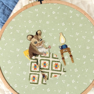 May include: Embroidered artwork in a wooden hoop. The design depicts a brown and white mouse in bed under a patterned blanket, with a small table and lamp. The background is a light green fabric with white dots.