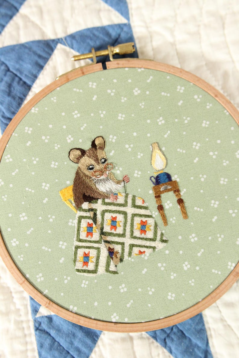 May include: Embroidered artwork in a wooden hoop. The design depicts a brown and white mouse in bed under a patterned blanket, with a small table and lamp. The background is a light green fabric with white dots.
