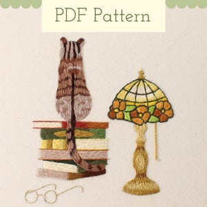 May include: Embroidered design of a cat sitting on a stack of books, a Tiffany-style lamp with orange flowers, and a pair of glasses. The design is on a beige fabric background.