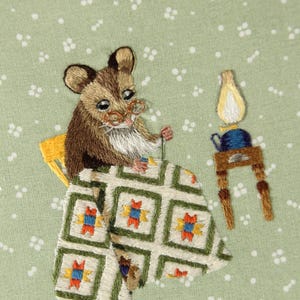 May include: Embroidered artwork showing a brown mouse wearing spectacles, seated at a table. The mouse is working on a quilt with a green, white, orange, and blue pattern. A small lamp and teapot sit on a table nearby. The background is a light green fabric.