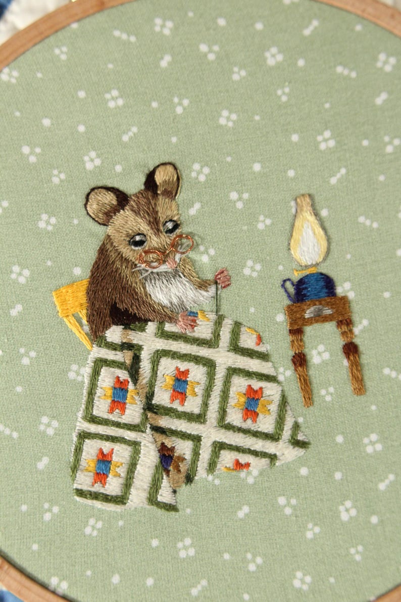 May include: Embroidered artwork showing a brown mouse wearing spectacles, seated at a table. The mouse is working on a quilt with a green, white, orange, and blue pattern. A small lamp and teapot sit on a table nearby. The background is a light green fabric.