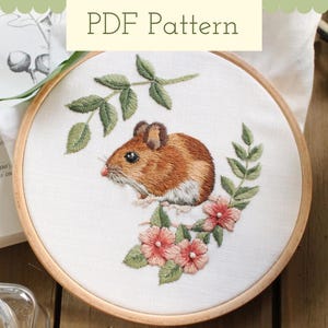 以下が含まれることがあります： ピンクの花と緑の葉が付いた茶色のネズミを刺繍したフープアート。ネズミは枝に座っており、尻尾は枝に巻き付いている。花は咲いており、葉は緑色。フープは木でできており、白い布の背景がある。
