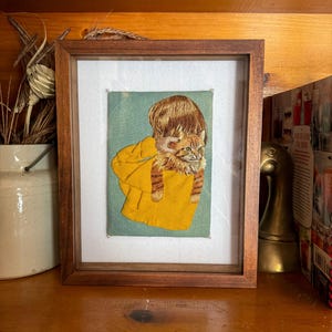 May include: A framed embroidery artwork featuring a ginger tabby cat peeking out from a bright yellow fabric pouch against a muted sage green background. The cat has detailed orange fur, green eyes, and is centered within a dark wooden frame.