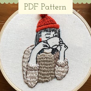 Può includere: Arte ricamata su un cerchio con una persona che indossa un cappello di lana rosso e un maglione marrone, tenendo una tazza di caffè o tè.