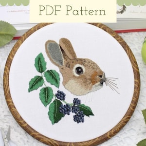 May include: Embroidered hoop art featuring a brown rabbit with black eyes and a white nose, surrounded by green leaves and black berries. The hoop is made of wood with a brown stain.