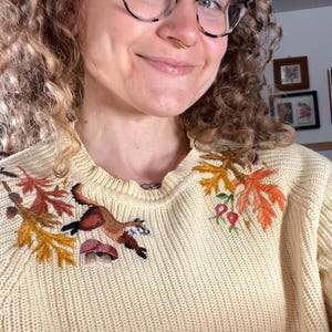 person with curly hair and glasses wearing a light yellow knit sweater with fox, and leaves, and mushrooms embroidered along the shoulders
