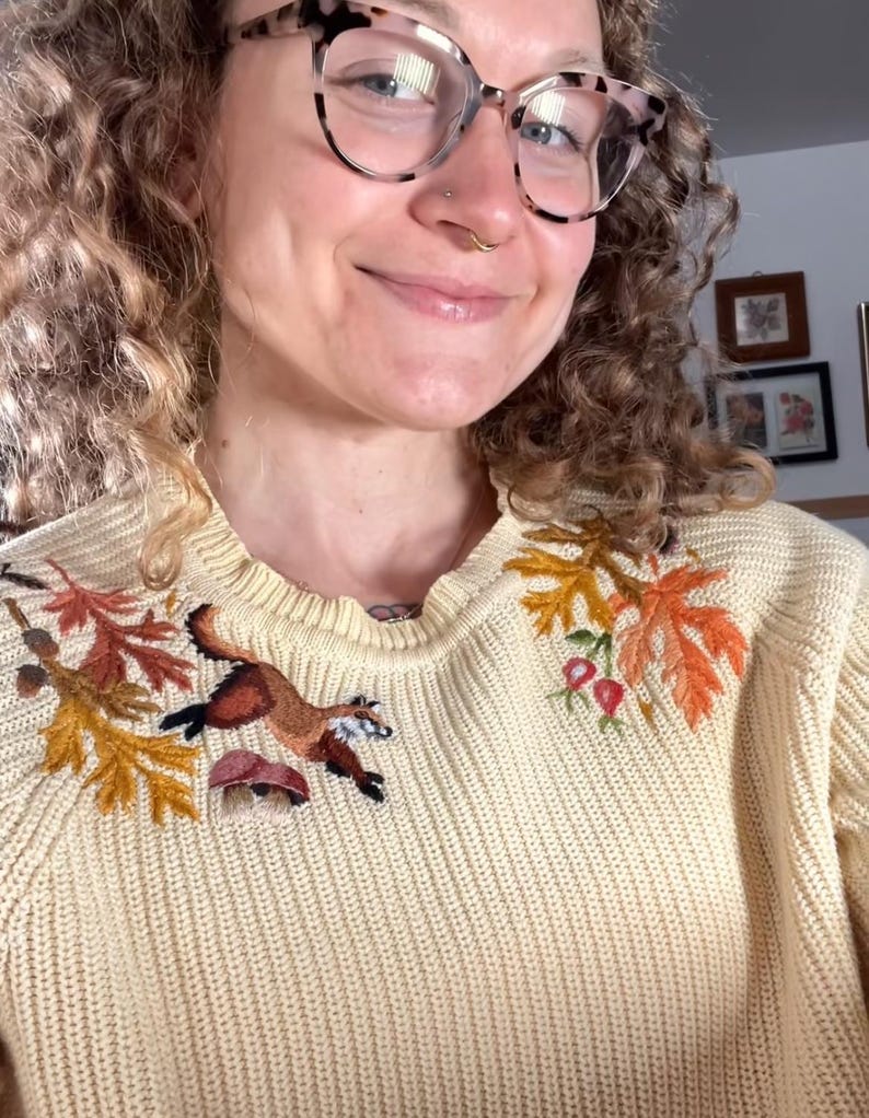 person with curly hair and glasses wearing a light yellow knit sweater with fox, and leaves, and mushrooms embroidered along the shoulders