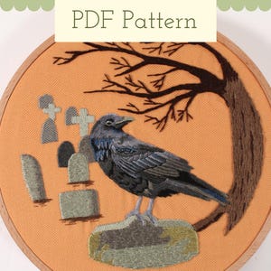 May include: Embroidered hoop art featuring a black crow perched on a rock in a graveyard setting. The crow is surrounded by gray tombstones and a brown tree branch.