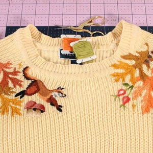 a yellow knit sweater with an embroidery along the neckline. The embroidery features a fox leaping over mushrooms, with colorful leaves, acorns, and rose hips.
