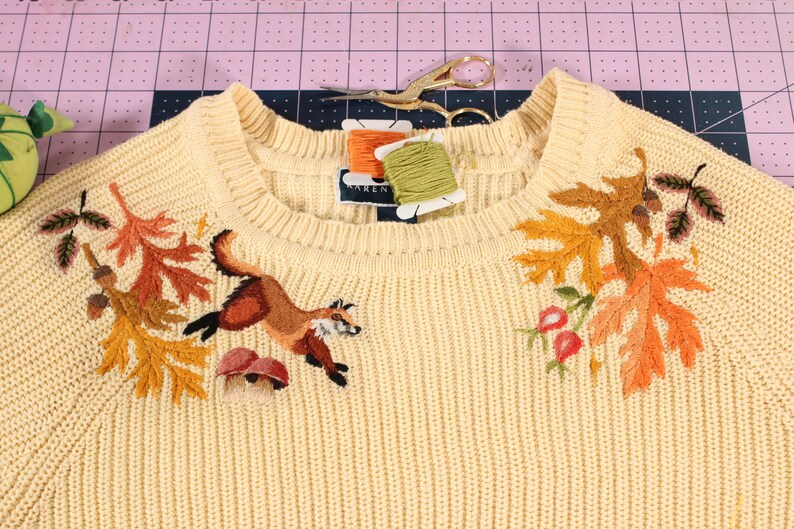a yellow knit sweater with an embroidery along the neckline. The embroidery features a fox leaping over mushrooms, with colorful leaves, acorns, and rose hips.
