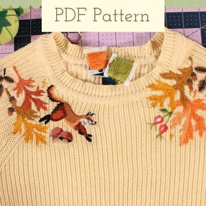 Puede incluir: Un suéter de punto color crema con una escena otoñal bordada. El diseño presenta un zorro, hojas, bellotas y setas en tonos naranja, marrón y verde.