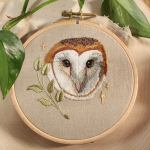 May include: Embroidered owl face in a wooden hoop. The owl has brown, white, and tan feathers, with a sprig of green leaves. The background is a tan fabric, with gold stars embroidered around the owl.