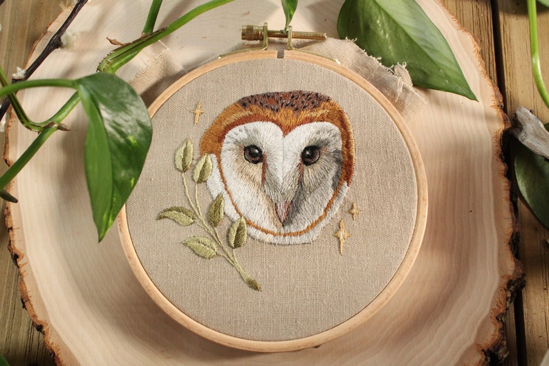 May include: Embroidered owl face in a wooden hoop. The owl has brown, white, and tan feathers, with a sprig of green leaves. The background is a tan fabric, with gold stars embroidered around the owl.