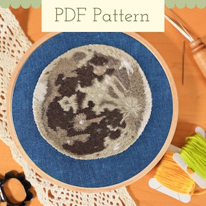 May include: An embroidery hoop with a detailed embroidered moon design on blue fabric. The text "PDF Pattern" is at the top. Spools of yellow and green thread, a needle, and lace are also visible, suggesting a craft project.