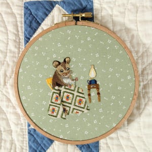 May include: Embroidered hoop art depicting a mouse at a table with a lamp. The mouse is brown and white, and the table has a patterned cloth. The background is a light green fabric with white dots.