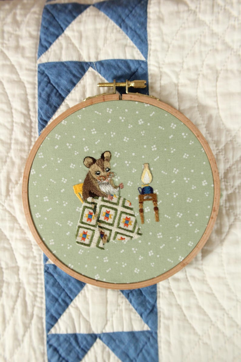 May include: Embroidered hoop art depicting a mouse at a table with a lamp. The mouse is brown and white, and the table has a patterned cloth. The background is a light green fabric with white dots.