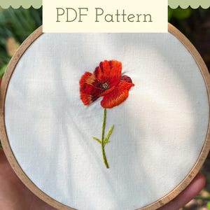 May include: A circular wooden embroidery hoop containing white fabric with a hand-stitched red poppy flower featuring orange and dark red thread details and a green stem. The text PDF Pattern is displayed in a light-colored box at the top.