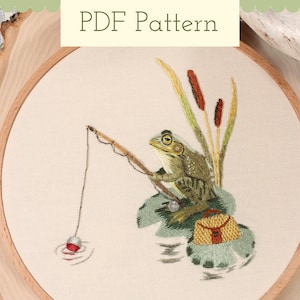 May include: Embroidered design of a frog sitting on a lily pad and fishing with a rod and line. The frog is green with brown spots and has a small brown bag on its back. The lily pad is green with a brown stem. The background is a light beige fabric.