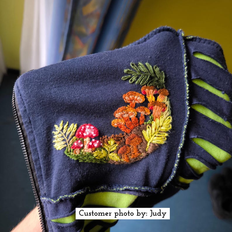 May include: Navy blue zip-up hoodie with an embroidered mushroom design. The embroidery features red, orange, and brown mushrooms with green and yellow foliage. The hoodie has a neon green lining and a zipper closure.