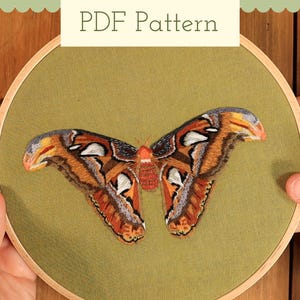 May include: A PDF pattern for an embroidery project featuring a detailed moth with orange, brown, white, and grey wings stitched onto olive green fabric. The embroidery is displayed within a circular wooden hoop, showcasing intricate thread work.