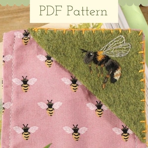 May include: A pink fabric bookmark with a green felt corner featuring an embroidered bee. The bookmark is decorated with a repeating pattern of small embroidered bees.