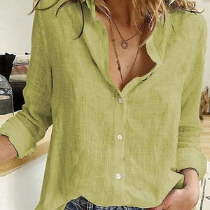 May include: A light olive green button-down shirt with a textured fabric finish, long sleeves, a classic collar, and a front button placket. The shirt is paired with blue denim jeans and layered necklaces, featuring a relaxed fit and visible weave.