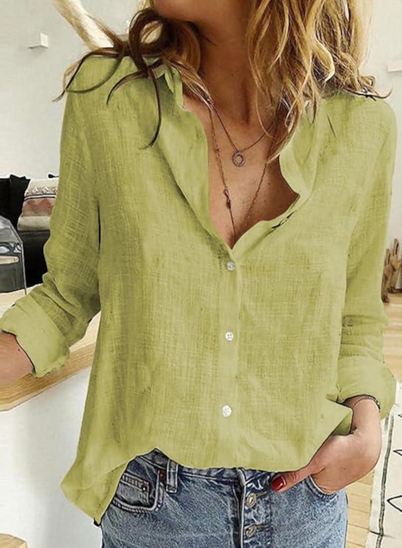 May include: A light olive green button-down shirt with a textured fabric finish, long sleeves, a classic collar, and a front button placket. The shirt is paired with blue denim jeans and layered necklaces, featuring a relaxed fit and visible weave.