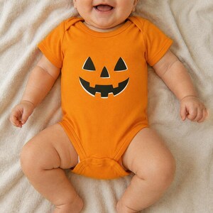 Puede incluir: Un body para bebé de color naranja brillante con un gráfico de cara de calabaza de Halloween en negro en el pecho. La prenda tiene mangas cortas, cuello redondo y cierre de botones a presión en la parte inferior para los bebés.