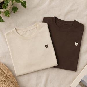 Puede incluir: Una camiseta color crema y una camiseta marrón oscuro, ambas con un pequeño corazón bordado en el pecho izquierdo. La camiseta crema tiene un corazón marrón oscuro con la letra A, mientras que la marrón tiene un corazón crema con la letra A.