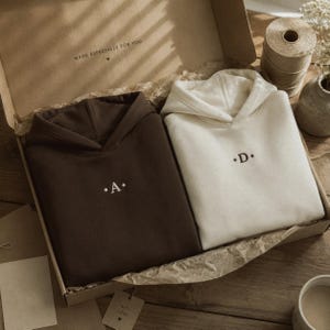 Puede incluir: Dos sudaderas con capucha empaquetadas en una caja de regalo de cartón. Una sudadera es de color marrón oscuro con una letra A bordada en blanco flanqueada por dos puntos. La otra es crema con una letra D bordada en marrón oscuro.