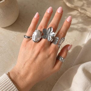 May include: A collection of five silver-toned rings worn on the fingers. The set includes a smooth band, a ring with a large white oval stone, a flower-shaped ring, a textured rectangular ring, and a curved ring with a perforated pattern.