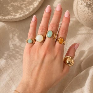 Könnte beinhalten: Eine Sammlung von fünf goldfarbenen Ringen an den Fingern. Vier Ringe haben glatte, ovale Cabochon-Steine in den Farben Hellblau, Weiß, Türkis und Bernstein. Der fünfte Ring ist ein breiter, goldfarbener Bandring mit gehämmertem Finish.