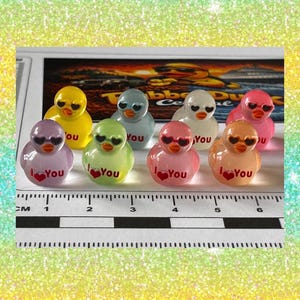 May include: Eight miniature rubber ducks in various colors including yellow, purple, green, blue, white, pink, and orange. Each duck wears heart-shaped sunglasses and features the text I Love You with a red heart. A ruler shows a scale in cm.