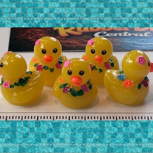May include: Four yellow rubber ducks with orange beaks, each decorated with small painted floral garlands in pink, blue, and green. The ducks are arranged on a white surface next to a ruler, with a sign reading Rubber Duck Central in the background.
