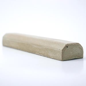 May include: A long, semi-cylindrical concrete object with a flat base and a smooth, light gray, porous surface. The piece features a beveled edge along the top length and a small, circular indentation on the flat end face, showing a solid texture.