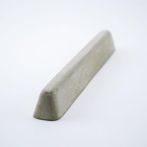 May include: A long, narrow, wedge-shaped object made of light gray concrete. The item features a smooth, matte finish with a slightly textured surface, showcasing a tapered profile that rises to a rounded top edge and a flat, stable base.
