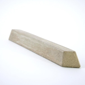 May include: A long, narrow, beige-colored object with a trapezoidal cross-section and a textured, porous surface. The item has a flat top, angled sides, and a flat base, appearing as a solid, elongated block with a matte, earthy finish.
